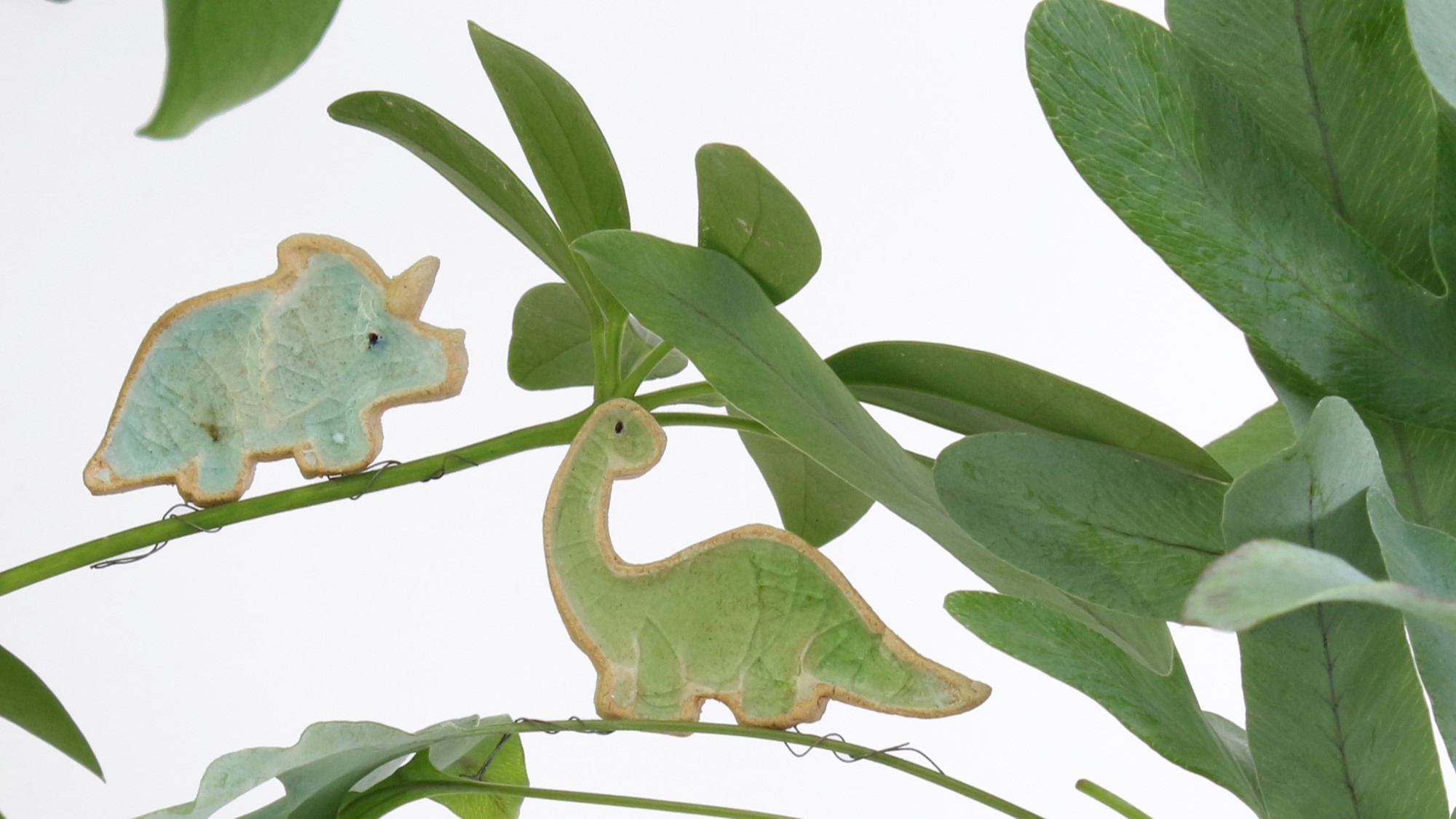 Two ceramic dinosaur figurines perched on a plant. One is a blue triceratops and the other is a green brontosaurus.