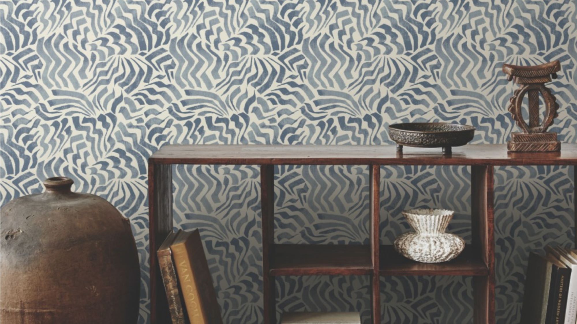 The image shows a room with a feature wall covered in a blue and white patterned wallpaper. The pattern is abstract and wavy, resembling stylised flowers or clouds. The wall is complemented by a wooden console table holding a collection of books, a vase, and a decorative bowl. A large ceramic jar stands to the left of the table, adding to the room's eclectic and cosy atmosphere.