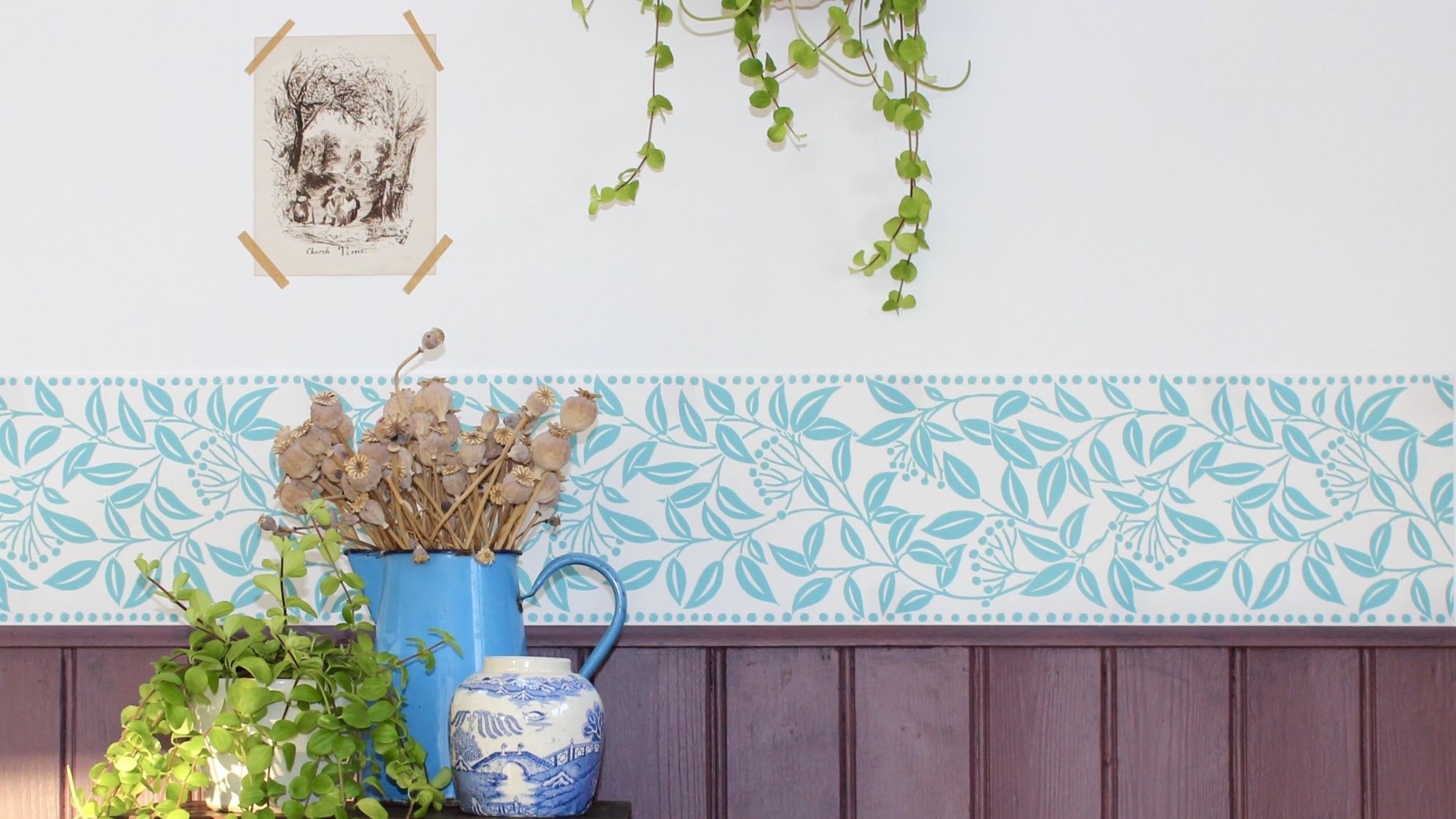 A room with a white wall and a wooden panelling at the bottom. There is a blue and white patterned border between the two. A picture frame with a black and white print hangs above the border. A vase with dried flowers sits on a table next to a small potted plant. A trailing plant hangs from a pot above the vase.