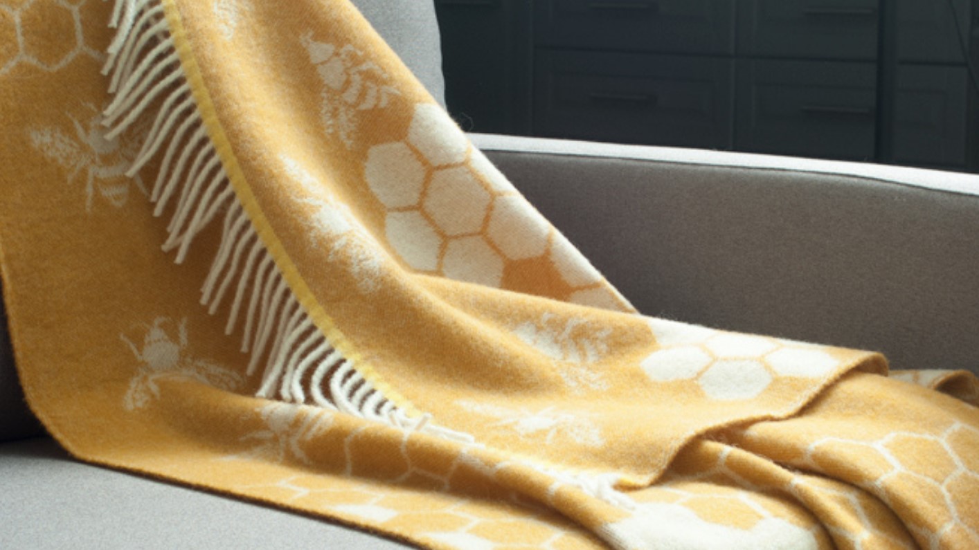 The throw features a distinctive pattern of bees and honeycombs, woven into a soft, mustard yellow wool. The fringes add a touch of texture and style. It seems to be a cozy and visually appealing piece that would complement a variety of home decor styles.