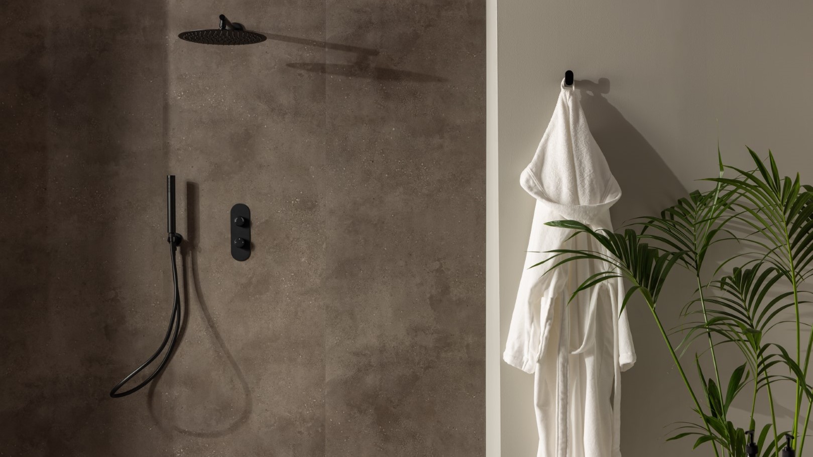 A modern bathroom with dark grey textured panel on the wall. A black rain shower head and matching thermostatic mixer valve are mounted on the wall. A white bathrobe hangs on a hook, and a large palm tree is in a corner.