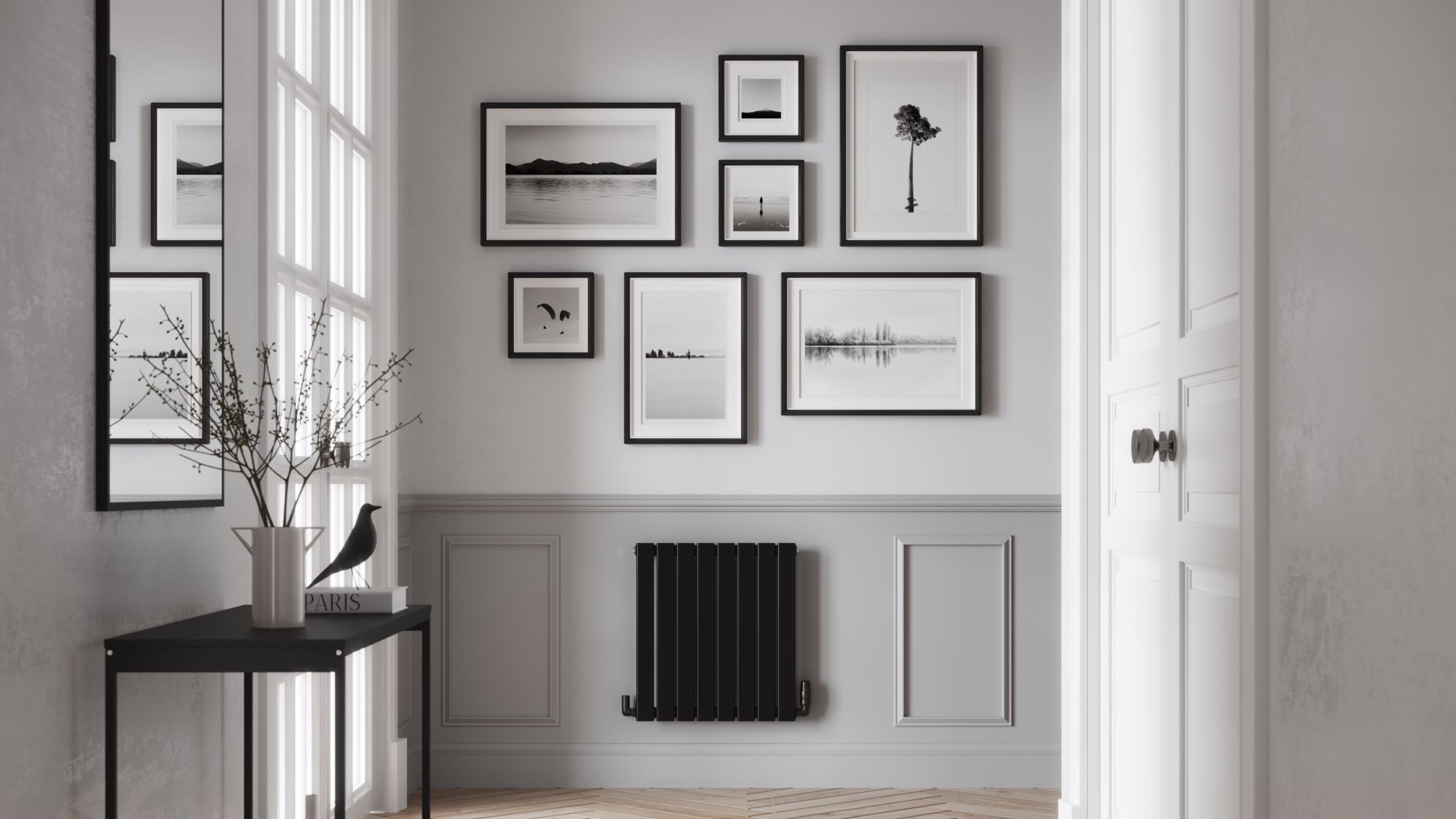 The images shows a single panel horizontal radiator with a matte black finish. It is positioned below a gallery wall of framed photographs. The hallway has grey walls, a wooden floor, and a door leading to another room.