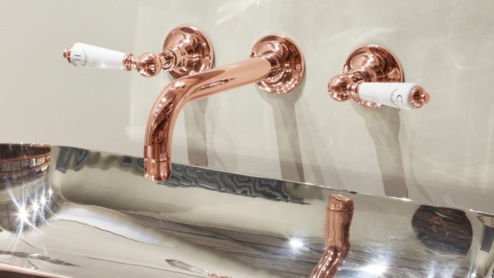 A luxurious bathroom vanity with a copper-coloured tap set. The tap has a classic design with white porcelain handles. The basin is rectangular and made of polished stainless steel. The overall aesthetic of the bathroom is elegant and sophisticated.