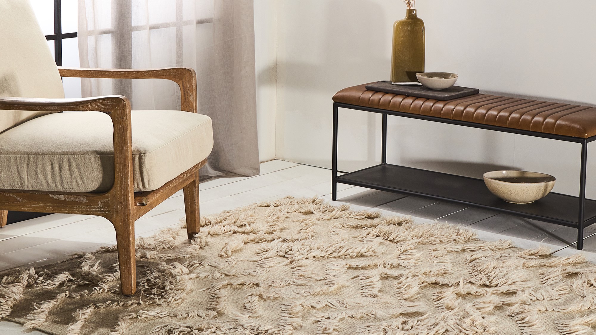 A living room with a beige rug, a wooden armchair, a metal bench with a leather seat, and a vase with dried flowers. The overall style of the room is bohemian and cosy.