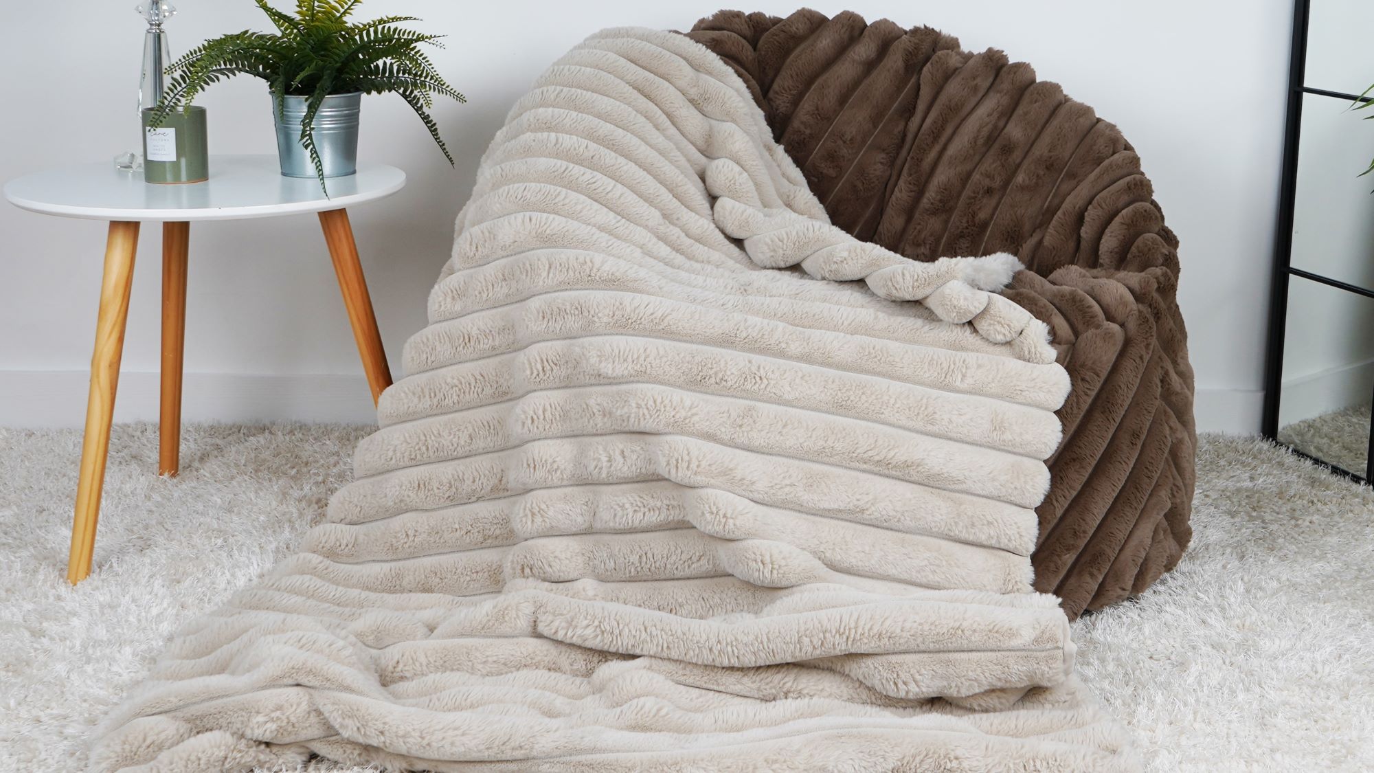 The image shows a faux fur throw blanket draped over a faux fur bean bag chair. The bean bag chair is a light brown colour, and the throw blanket is a light beige colour. The throw blanket has a ribbed texture and is very soft. The bean bag chair is placed in a living room setting, with a white coffee table and a potted plant nearby.