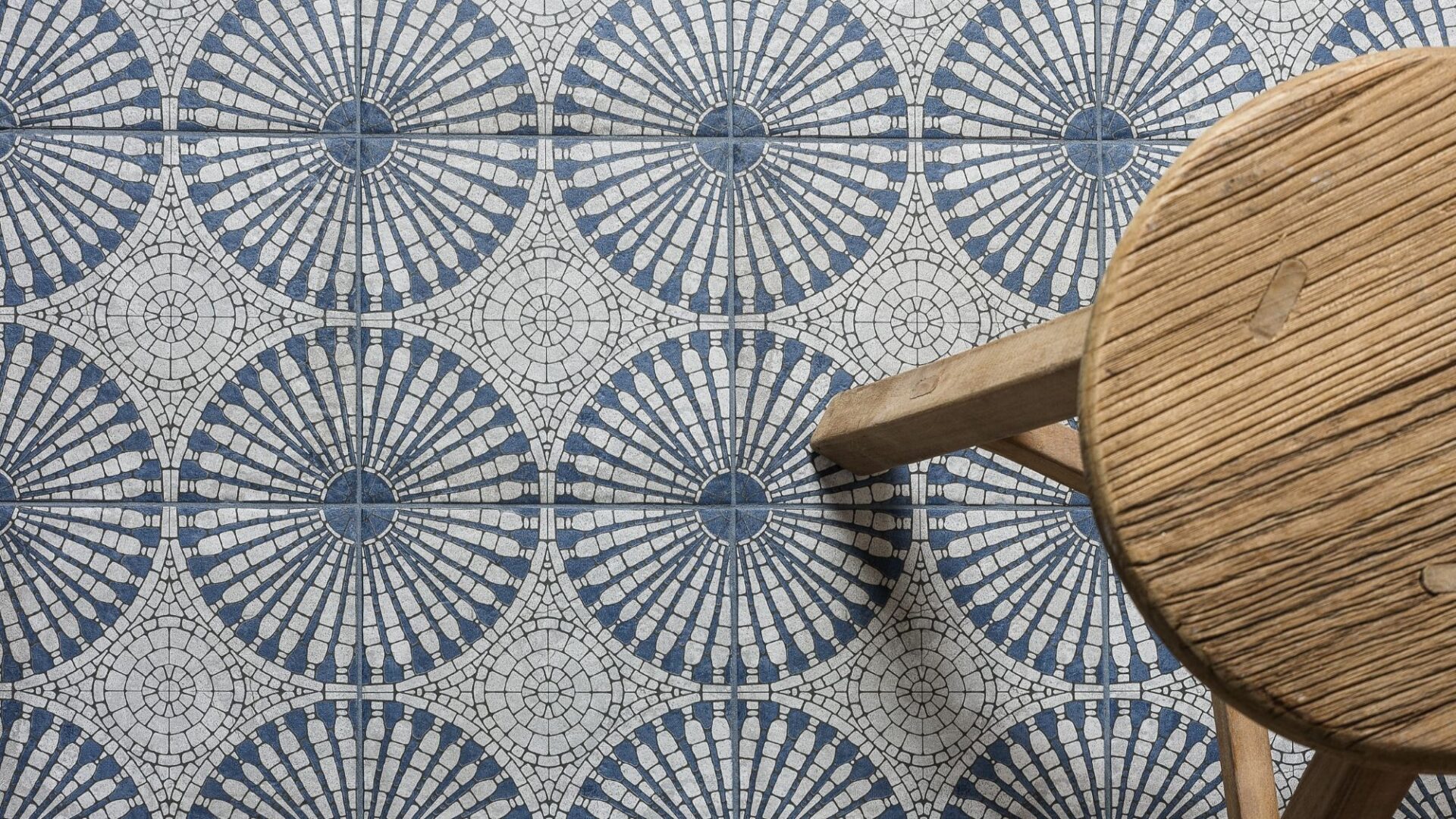 A bird’s eye view of a patterned floor tile. The light grey and blue tile has a circular geometric pattern. A small wooden stool is also seen.